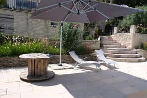 A patio area with a round wooden table, two white lounge chairs, and a large umbrella. - Vacation rental house with pool PUGET LS2-356 (Puget)