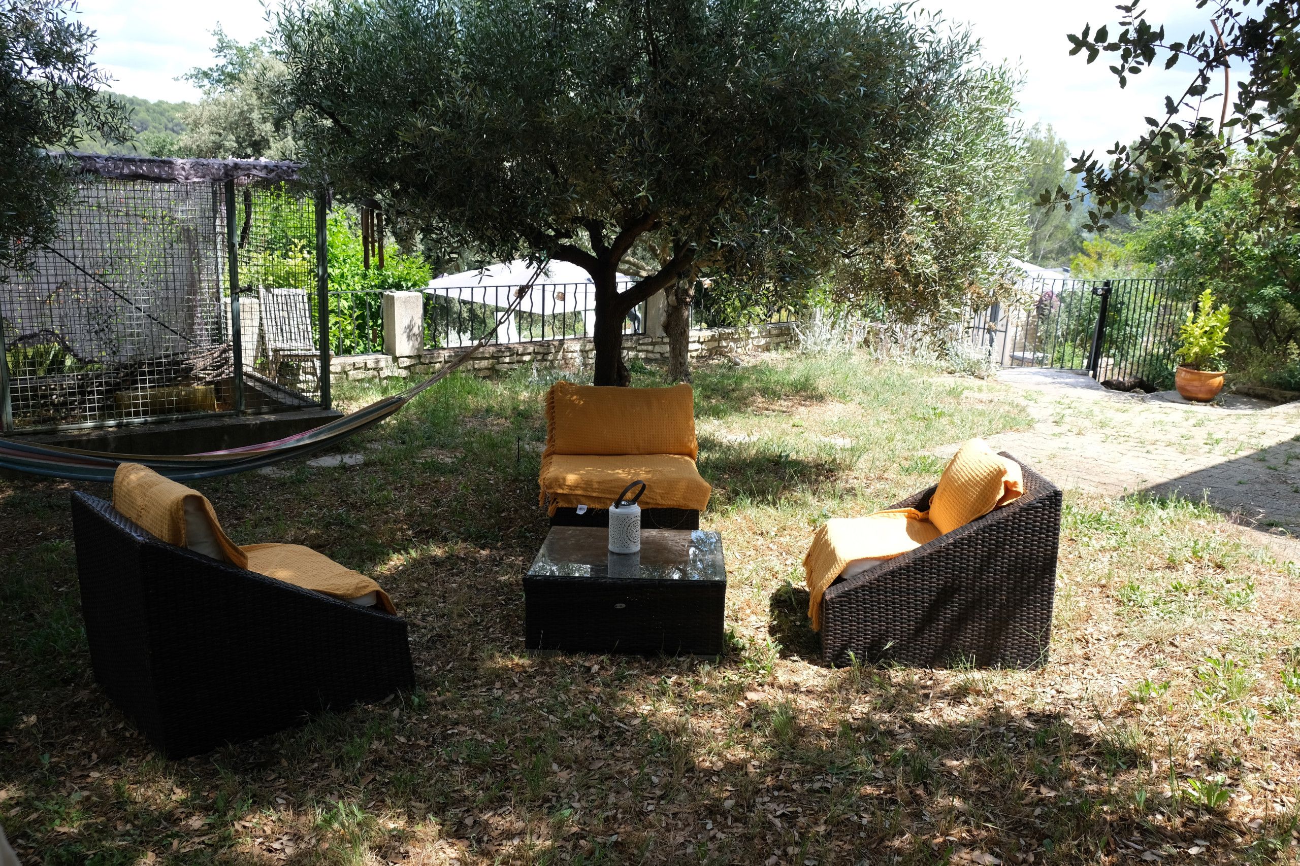 An outdoor seating area with wicker furniture and a small table, surrounded by greenery and a tree.