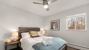 Un dormitorio con cama, dos mesitas de noche con lámparas, un ventilador de techo, una ventana con persianas y un cuadro en la pared.