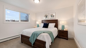 Um quarto com cama de madeira, duas mesas de cabeceira com abajures, uma janela com persianas e duas obras de arte emolduradas na parede.