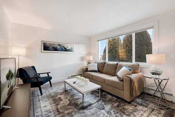 A modern living room with a sofa, coffee table, and a TV. There is a window with a view of trees and a painting on the wall.