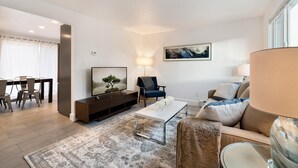 Uma sala de estar moderna com televisão de tela plana, um sofá, uma mesa de centro e um espaço de jantar com cadeiras.
