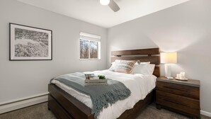 A bedroom with a wooden bed, a nightstand with a lamp, a framed picture on the wall, and a window with a view of trees.