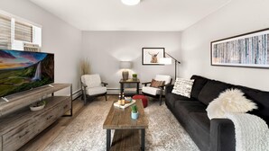 A modern living room with a flat-screen TV, a wooden coffee table, a black sofa, and a white armchair.