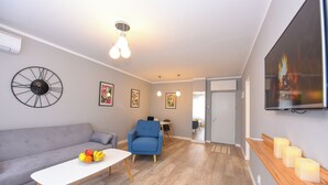 Een moderne woonkamer met een grijze bank, een blauwe fauteuil, een witte salontafel met fruit, en een televisie aan de muur.