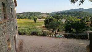 Une vue depuis un bâtiment en pierre donnant sur une zone de gravier, avec une clôture, des arbres et des collines au loin.