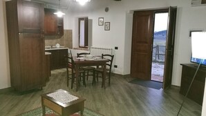 Un coin repas avec une table et des chaises, une petite table ornée d’un chemin de table, un espace cuisine avec des placards, et une vue à travers une porte ouverte.