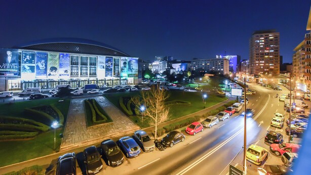Een nachtzicht van een stadsbeeld met een groot, verlicht gebouw, een parkeerplaats met auto's, en omliggende stadsgebouwen.
