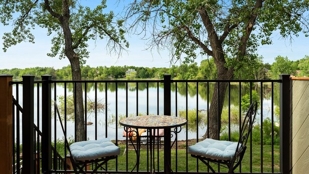 Terraza con mesa y dos sillas, con vistas a un lago con árboles y casas al fondo.