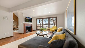 Una sala de estar moderna con chimenea, un sofá, un sillón amarillo y un ventanal con vistas a árboles y un lago.