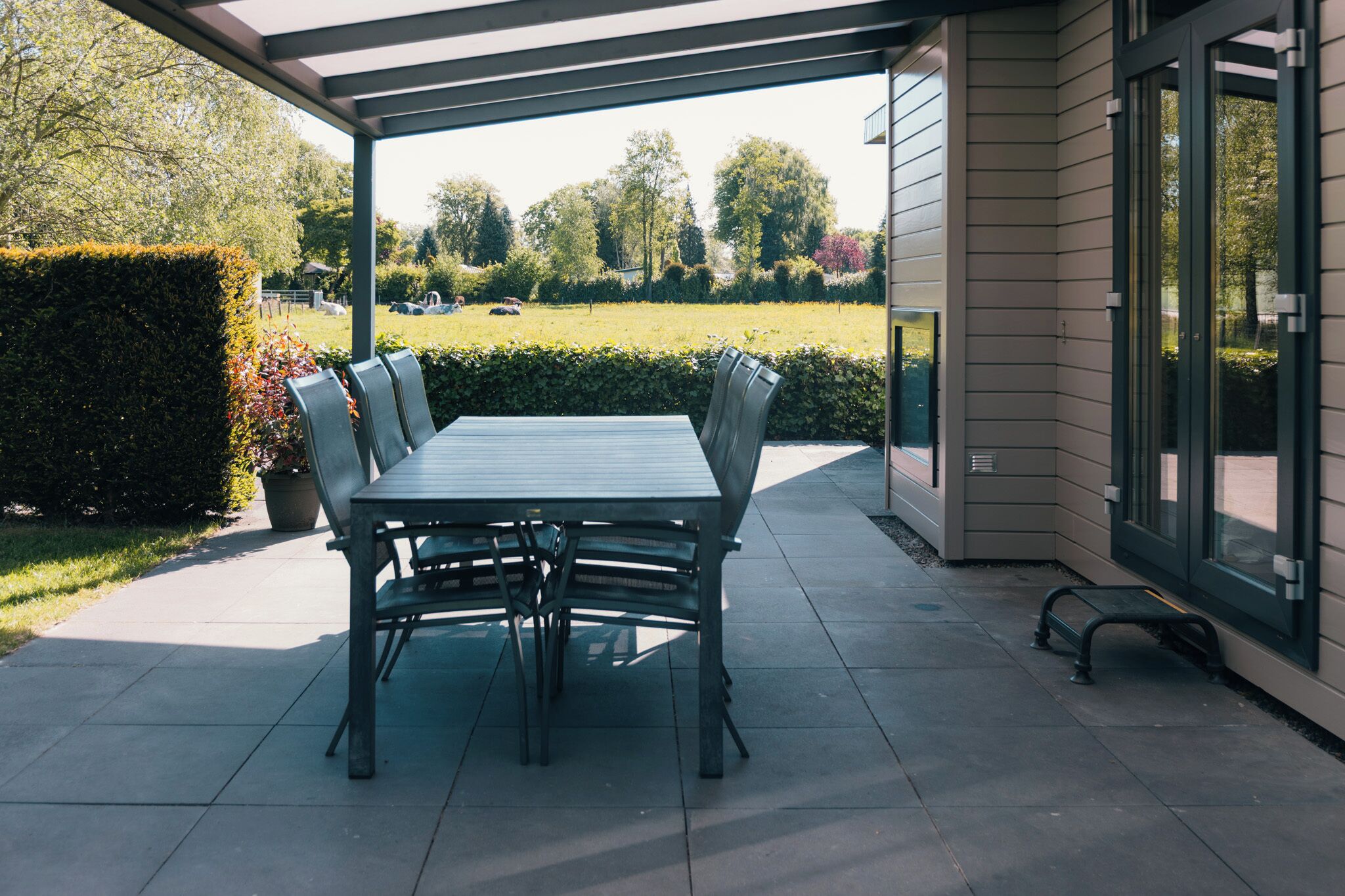 Eine Terrasse im Freien mit Tisch und Stühlen, einem überdachten Bereich und Blick auf Garten und Bäume.