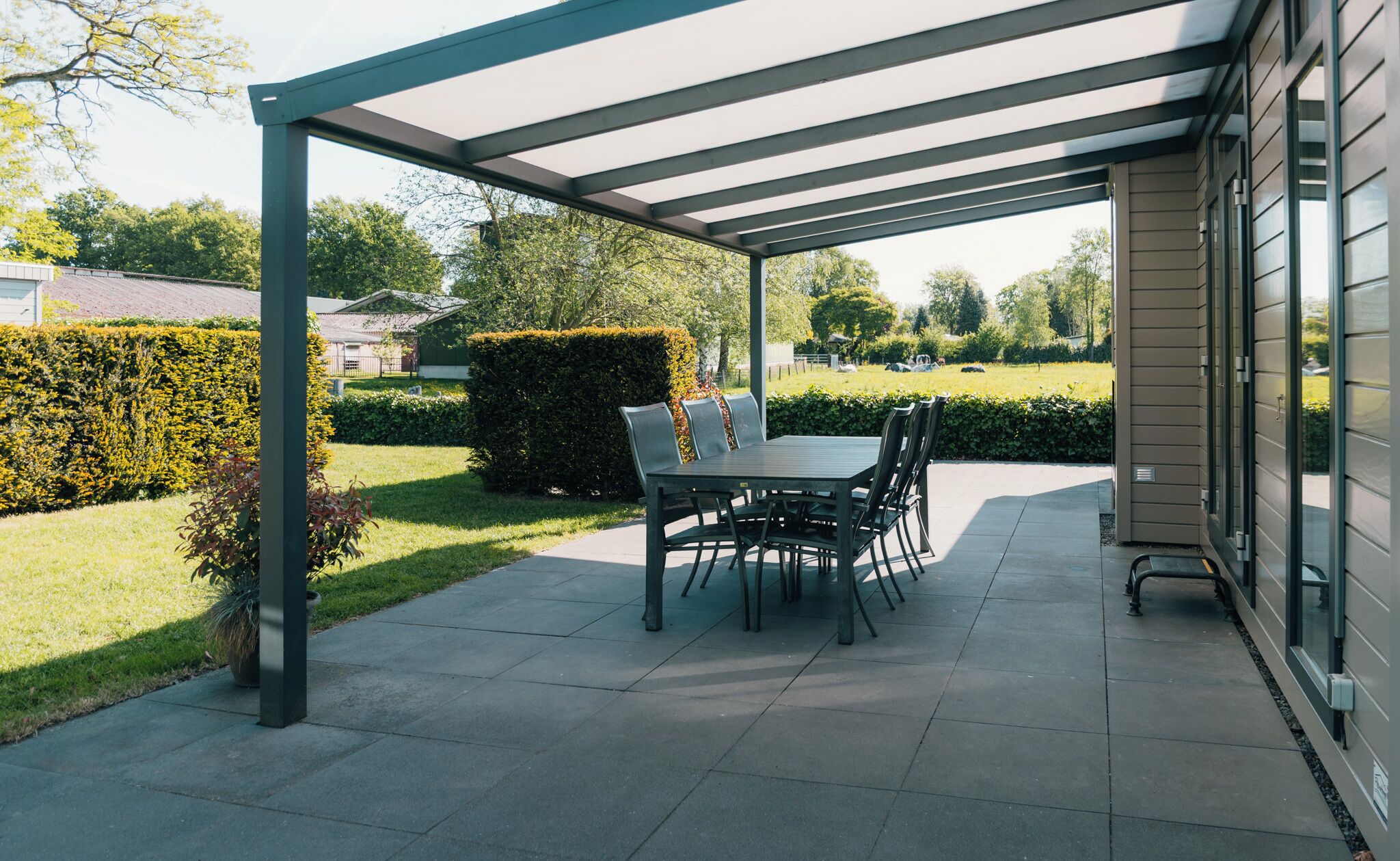 Un patio couvert avec une table et des chaises, entouré d’un jardin bien entretenu et de haies.