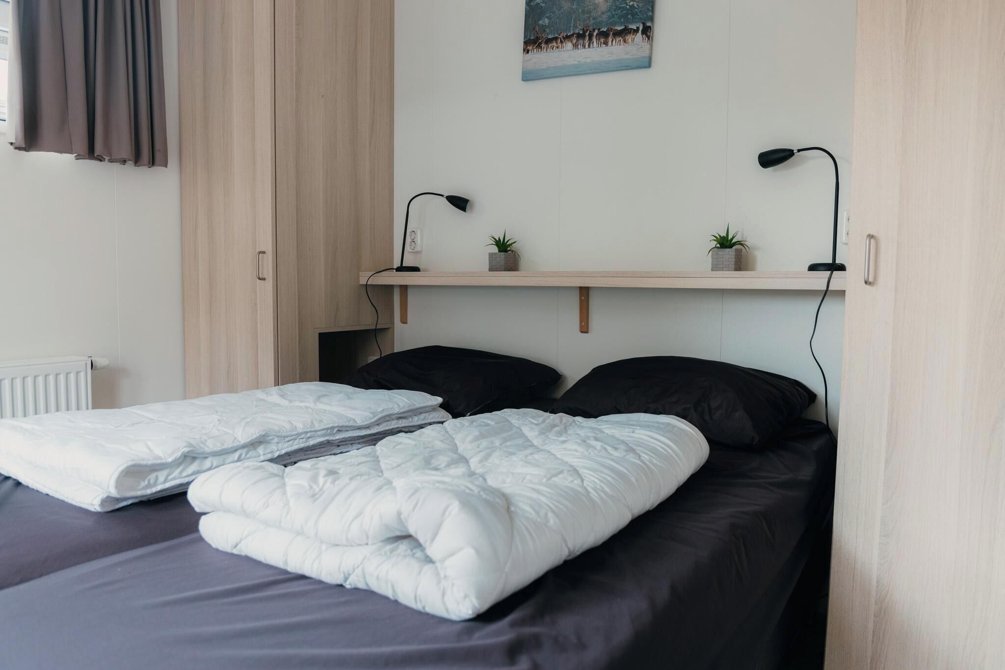 Een netjes opgemaakt bed met zwart-witte beddengoed, een plank met twee planten in potten, en een schilderij aan de muur.
