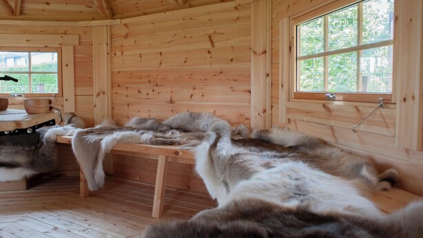 A wooden cabin interior with a fur-lined bench, wooden walls, and a window with a view of greenery.