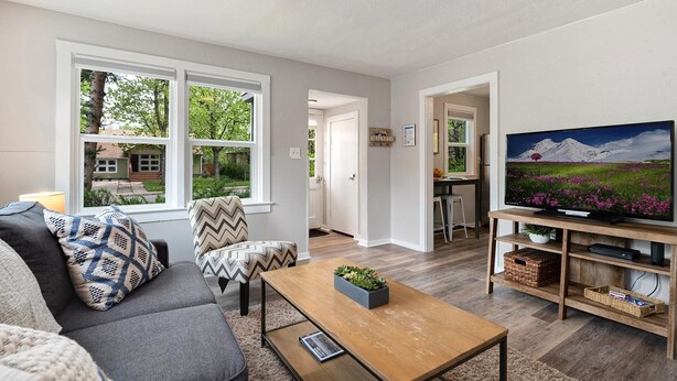 A living room with a sofa, coffee table, and TV. There are windows with a view of greenery and a door leading to another room. - Charming Old Town Bungalow w/ FREE Cruiser Bikes (Fort Collins)