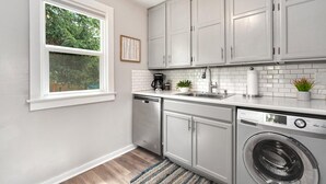 A compact kitchen with white cabinets, a dishwasher, a washing machine, a window with a view of greenery, and a small potted plant on the counter. - Charming Old Town Bungalow w/ FREE Cruiser Bikes (Fort Collins)