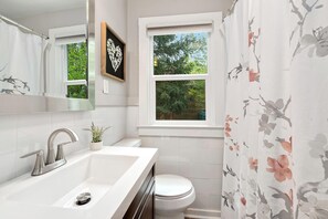 Et badeværelse med en hvid vask, toilet, et vindue med udsigt til grøn natur og et spejl med et hjerteformet kunstværk.