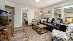 A modern living room with a sofa, coffee table, and a TV. There is a door leading to a bedroom and a window with a view. - Charming Old Town Bungalow w/ FREE Cruiser Bikes (Fort Collins)