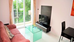 A living room with a sofa, a glass coffee table, a flat-screen TV, and a balcony with curtains.