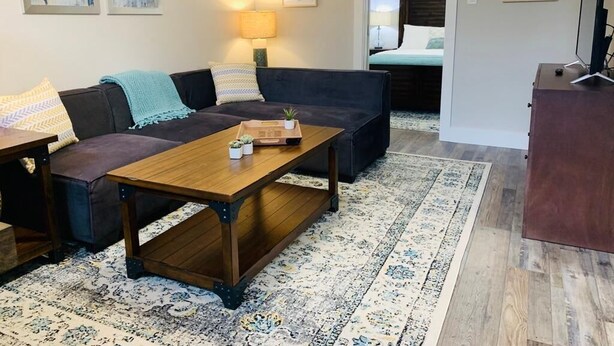 Sala de estar com um sofá escuro, uma mesa de centro de madeira e um tapete estampado. Há uma cama ao fundo e uma TV sobre um suporte.