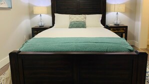 Quarto com cama de madeira escura, roupa de cama branca, manta verde, duas luminárias de cabeceira e uma janela com persianas.