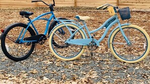 Uma bicicleta azul com cesto e bagageiro, estacionada sobre cascalho, com uma cerca de madeira ao fundo.