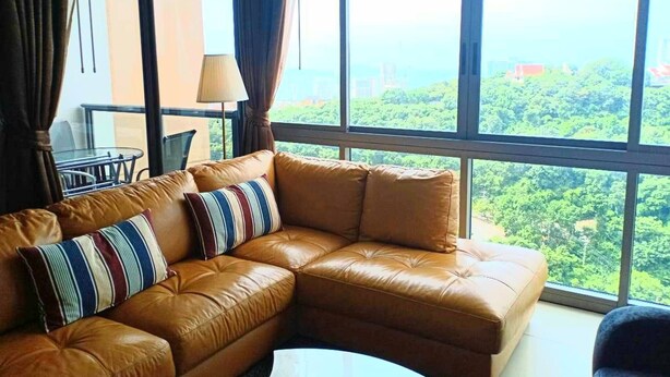 A brown leather sofa with striped pillows, a white table, and a view of greenery through large windows. - 3110 - 2 Bedroom Seaview Deluxe Apt Unixx Pattaya (Pattaya)
