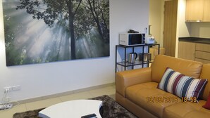A living room with a large forest-themed painting, a brown leather sofa, a coffee table, and a kitchen area in the background. - 3110 - 2 Bedroom Seaview Deluxe Apt Unixx Pattaya (Pattaya)