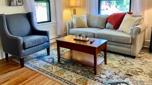 A living room with a dark armchair, a beige sofa, a wooden coffee table, and a patterned rug.