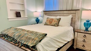 A neatly made bed with a white comforter and a colorful woven blanket, a wooden headboard, a nightstand with a blue lamp, and a wall-mounted shelf with a decorative item.