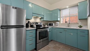 A kitchen with teal cabinets, stainless steel appliances, and a window with blinds.