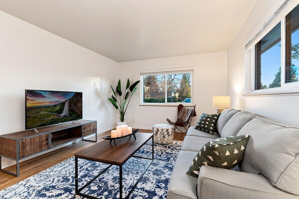 A modern living room with a flat-screen TV, wooden coffee table, and a grey sofa with patterned cushions.