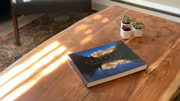 En bok med titeln ”Colorado – en historisk atlas” ligger på ett träbord tillsammans med två små krukväxter.