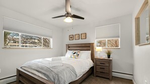 A bedroom with a wooden bed, a nightstand, a ceiling fan, and two windows with blinds. - Contemporary condo in the heart of Old Town (Fort Collins)