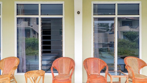 A covered outdoor seating area with wicker chairs and a glass table, adjacent to large windows with a view of a residential area. - OYO 3250 Setia Hotel (Cipanas)