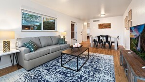 A modern living room with a grey sofa, a coffee table, and a patterned rug. There is a dining area with a table and chairs, and artwork on the walls.