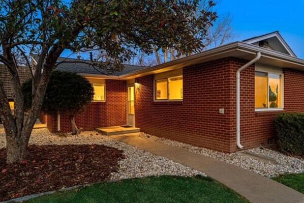 A two-story brick house with a front porch, a small tree, and a well-maintained lawn.