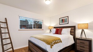 A bedroom with a wooden bed, a window with a view, a ladder, two bedside tables with lamps, and a framed picture on the wall.