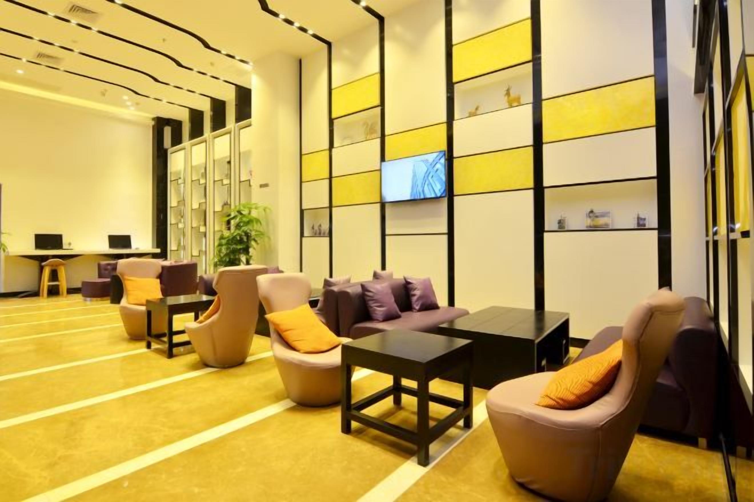A hotel lobby with a reception desk, seating area with armchairs and sofas, a television screen, and a wall with shelves.