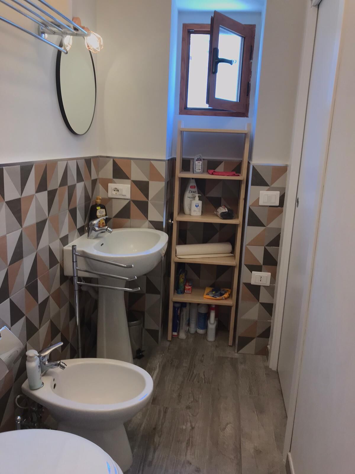 A compact bathroom with a toilet, bidet, and sink. There is a mirror above the sink, a shelf with toiletries, and a window above the door.