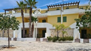 A courtyard with a stone floor, palm trees, and a building with balconies and green shutters. - Villetta Sole (Gallipoli)