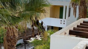 A courtyard with palm trees, a stone floor, and a white building with green shutters. - Villetta Sole (Gallipoli)