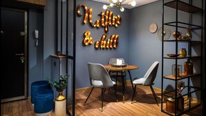Una camera moderna con un cartello luminoso con scritto "Netflix & chill", un tavolo rotondo in legno, due poltrone grigie, un poggiapiedi blu, una pianta in un vaso dorato e una scaffalatura in metallo nero.