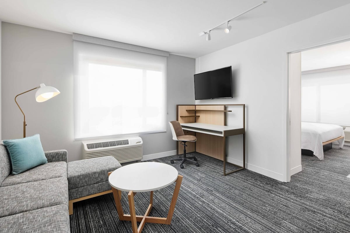 Moderni hotellihuone, jossa on harmaa sohva, pieni pyöreä pöytä, työpöytä tuolilla, taulutelevisio ja sänky.