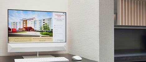 Una habitación de hotel con escritorio, monitor de computadora, teclado, ratón e impresora.