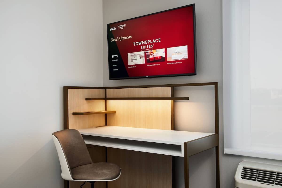 Moderni hotellihuone, jossa on työpöytä, tuoli ja televisio, jossa näkyy teksti: »Hyvää iltapäivää» ja »TOWNEPLACE SUITES».