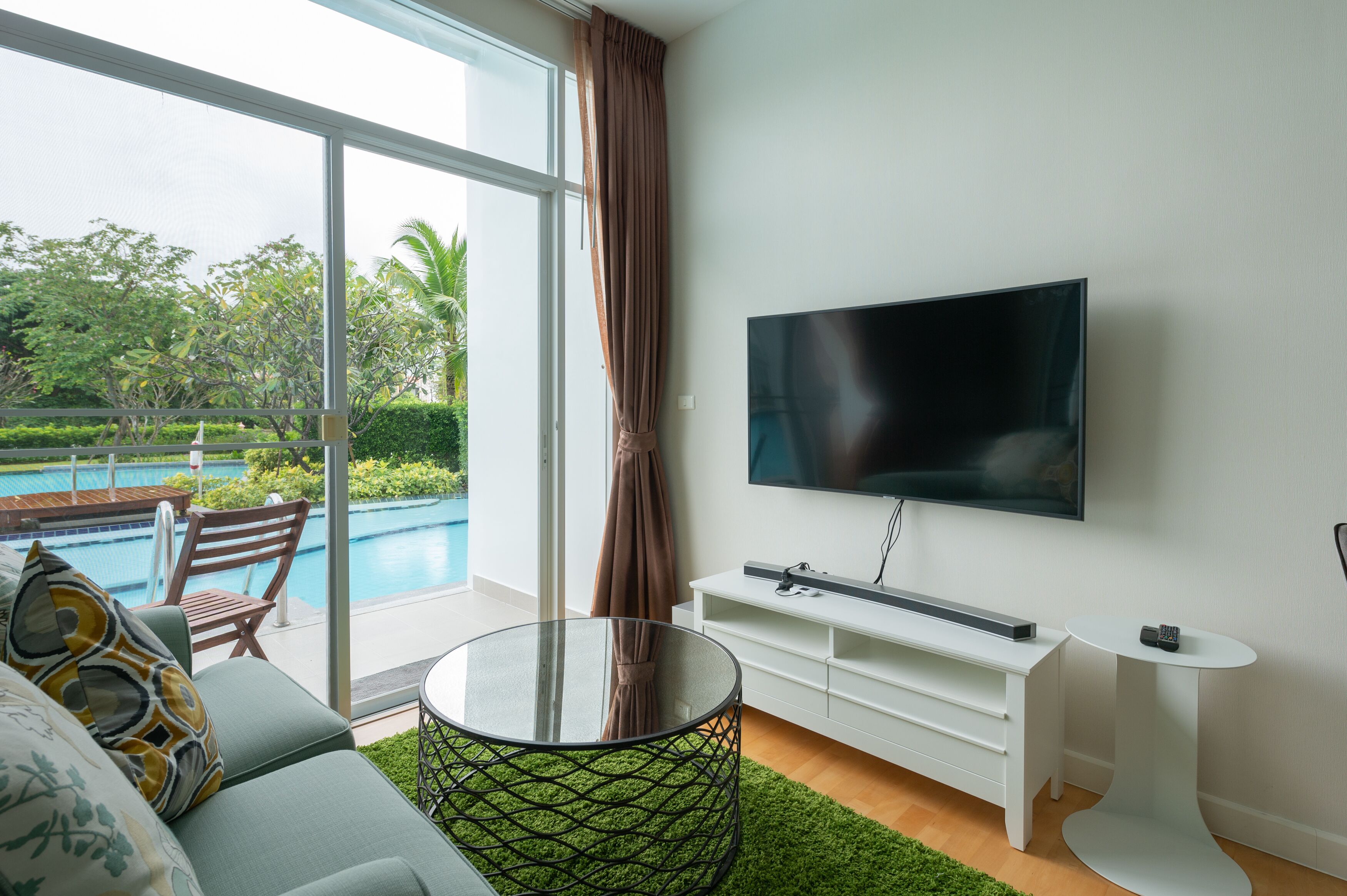 A modern living room with a large TV, a glass coffee table, a sofa, and a view of a swimming pool.