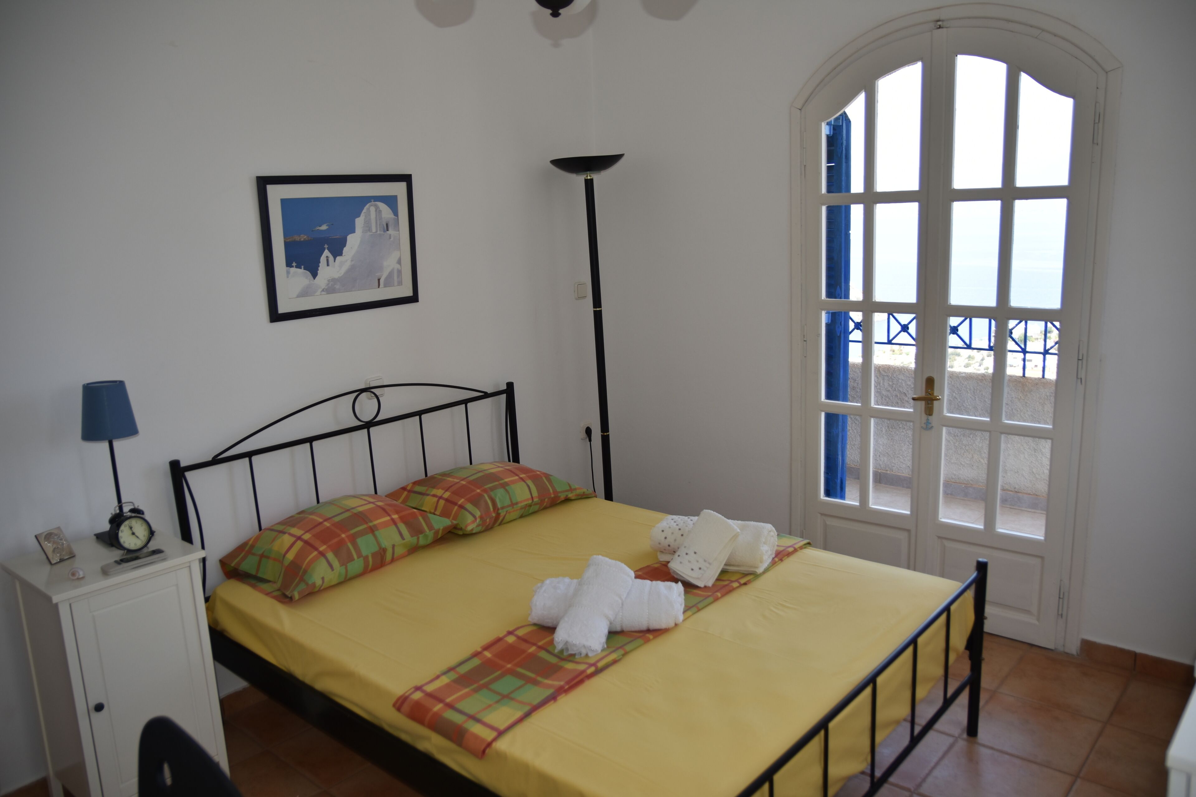 Cama bem arrumada com colcha amarela, manta xadrez e toalhas brancas. Há um quadro na parede, uma mesa de cabeceira com abajur e uma janela com vista para a praia.