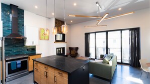 A modern kitchen with a large island, built-in oven, and a stone backsplash. There is a sofa and a dining area with a large window. - Broken Hill Designer Baker St Flat Mountain View.  (Nelson)