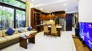 Un salón moderno con sofá, mesa de centro y televisor. Se ve una cocina con armarios de madera y una zona de comedor con sillas.